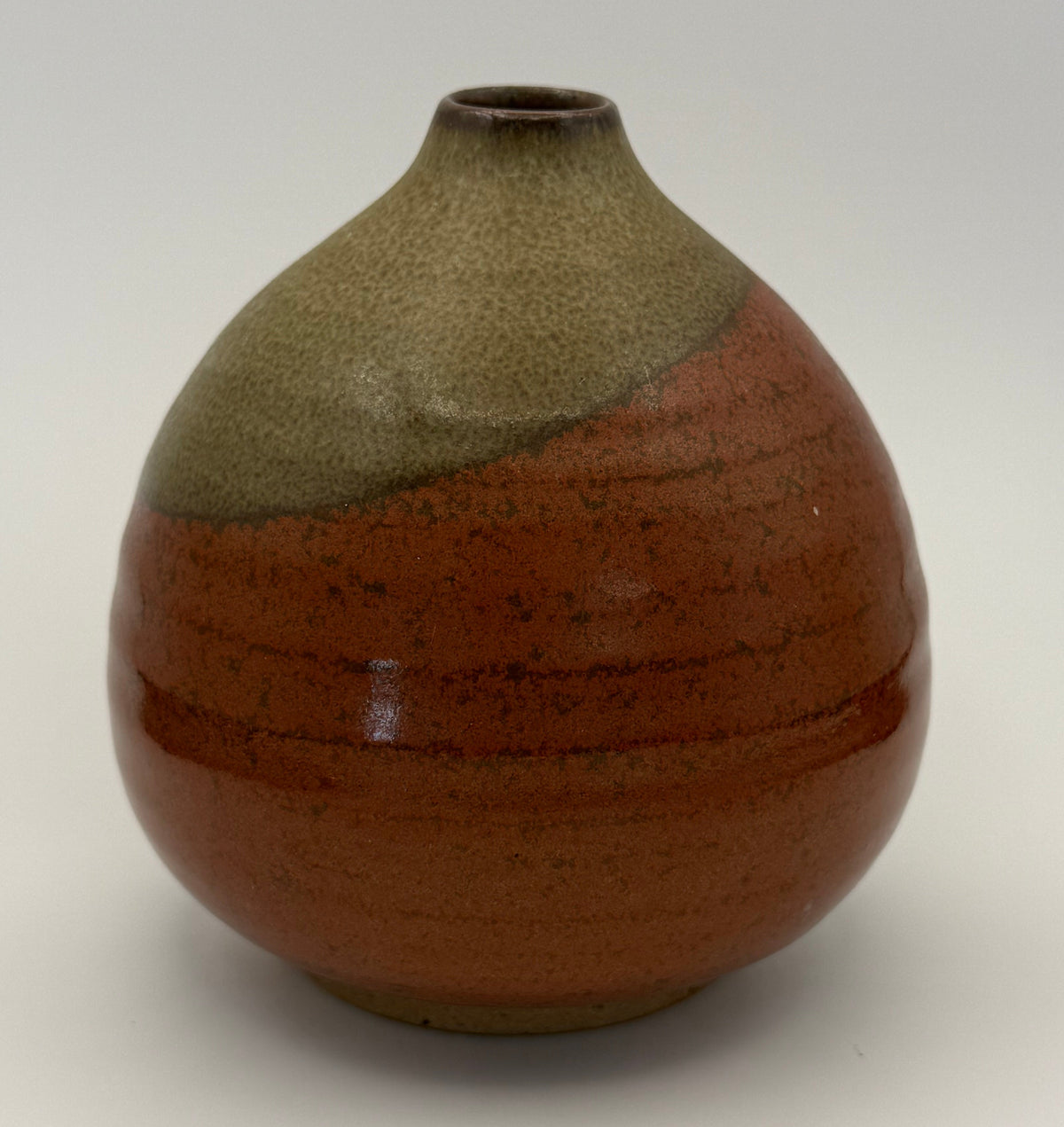 A handmade ceramic vase with a rounded body, a narrow neck, and a small flared top. It features a two-tone glaze: a textured brown glazed top half and a speckled reddish-brown glazed bottom half, set against a plain white background.