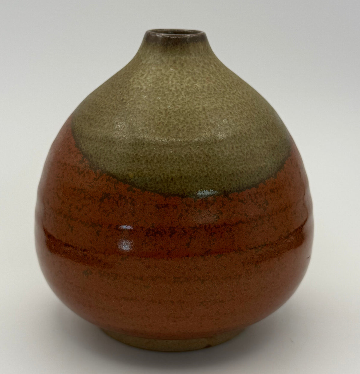 A handmade ceramic vase with a rounded body, a narrow neck, and a small opening at the top. It features a two-tone glaze: a textured brown top half and a glossy reddish-brown bottom half with subtle horizontal ridges, set against a white background.