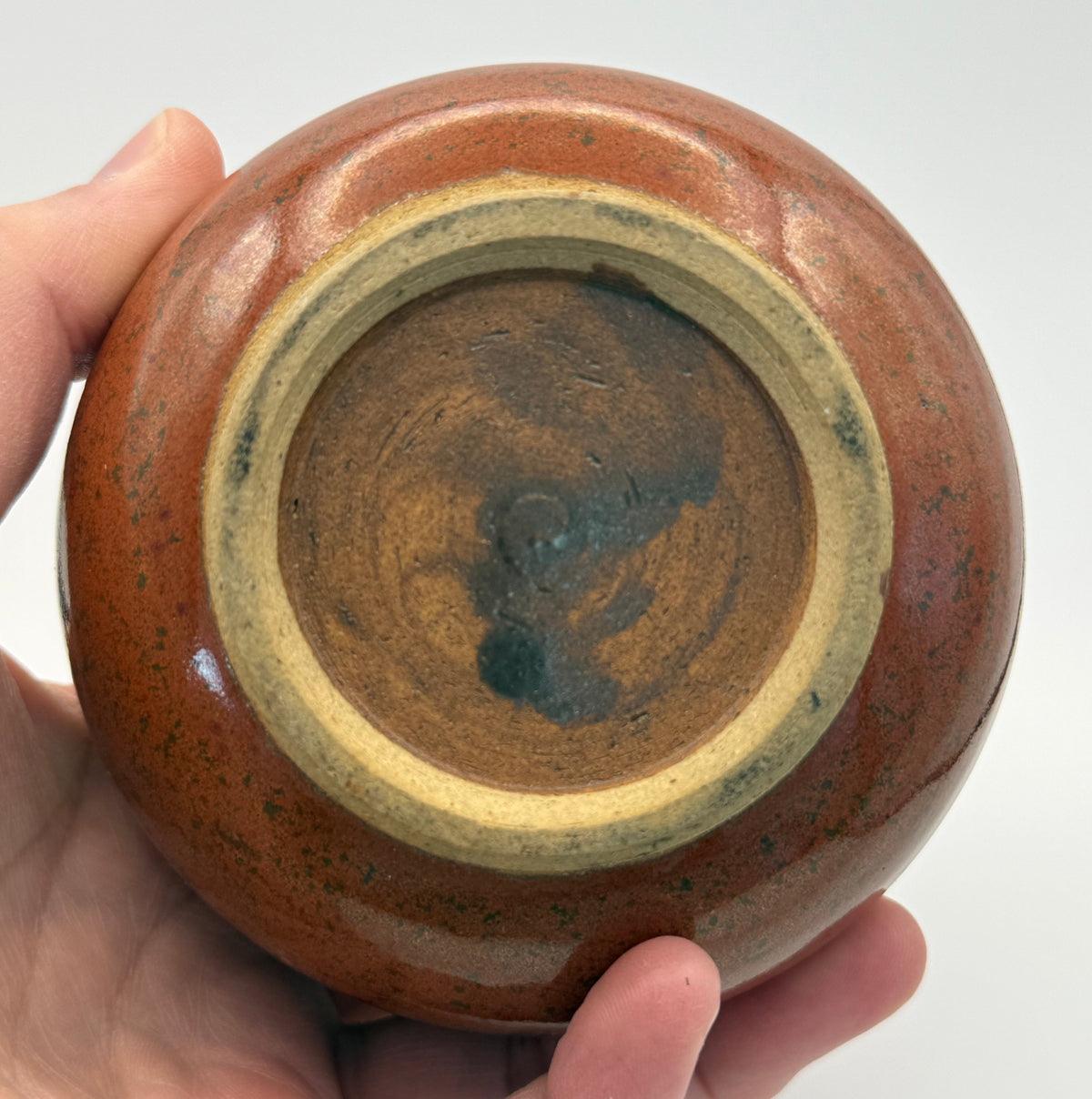 A hand holding the base of a small round ceramic vessel to show the glazed interior and unglazed clay rim against a plain light background.
