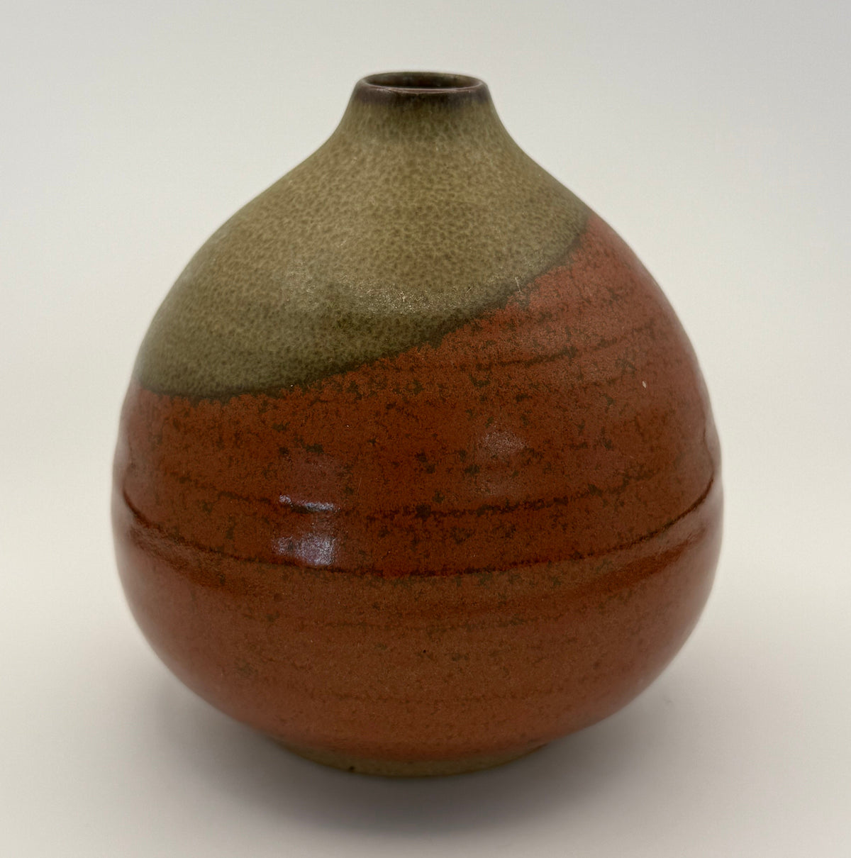 A handmade ceramic vase with a rounded bulbous body, a narrow neck, and a small opening at the top. It features a two-tone glaze: a speckled brown and orange-brown lower section and a matte olive-green upper section, set against a plain white background.