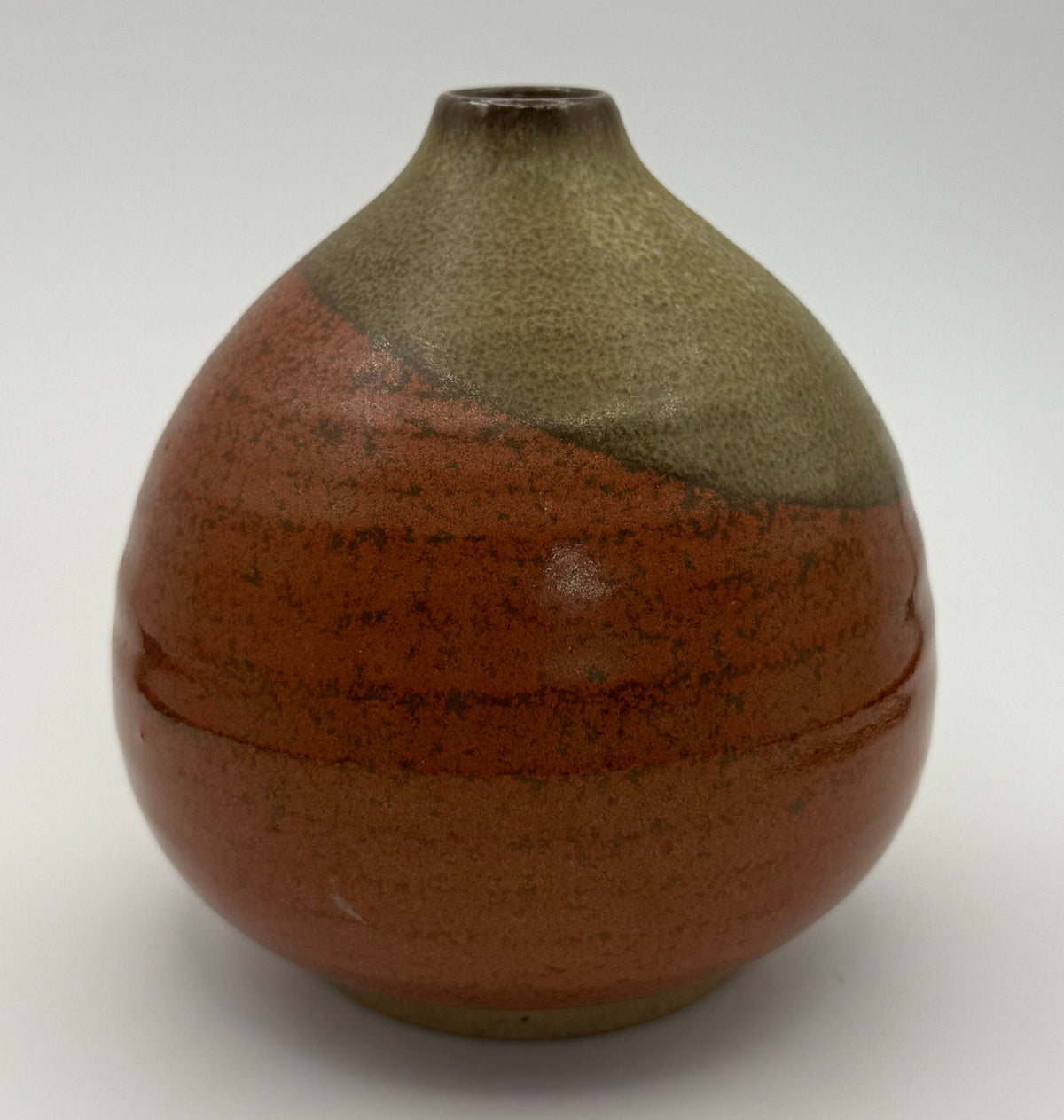 A handmade ceramic vase with a rounded bulbous body, a narrow neck, and a small opening at the top. It features a two-tone glaze: a textured reddish-brown lower section and a speckled brown upper section, set against a white background.
